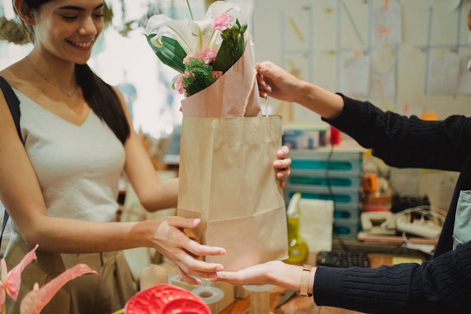 A woman wearing a white sleeveless top is smiling as she receives a bouquet arrangement of fresh flowers from a floral shop assistant. The bouquet features white calla lilies and pink roses, wrapped i