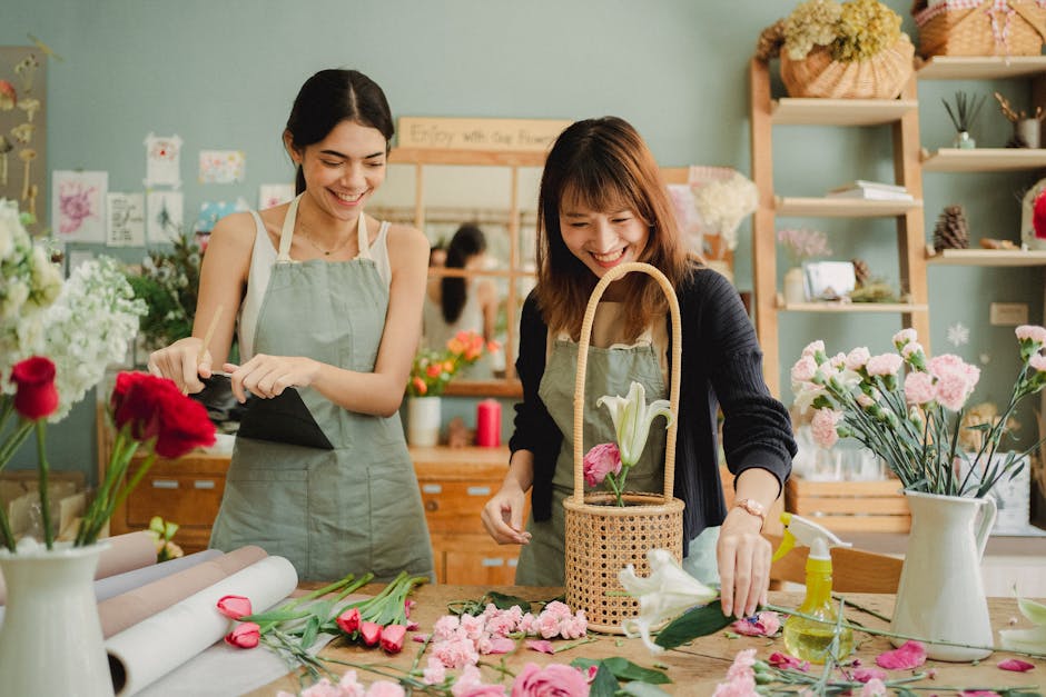 Two women in a flower shop, smiling and working together on floral arrangements. The woman on the left, wearing a light green apron, is holding a black wrapping paper, while the woman on the right, in