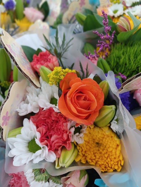 A vibrant floral arrangement featuring a central orange rose surrounded by white daisies, pink carnations, and small yellow flowers. The bouquet is wrapped in delicate, semi-transparent white paper wi