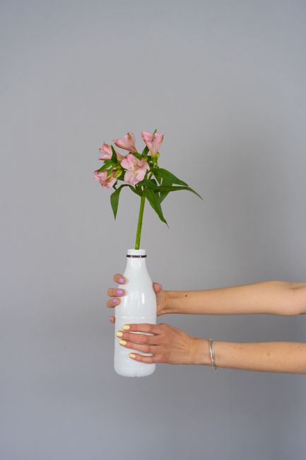 A person with well-manicured nails painted in pastel shades is holding a simple white ceramic vase containing a fresh floral arrangement of pale pink flowers with green leaves. The flowers are arrange