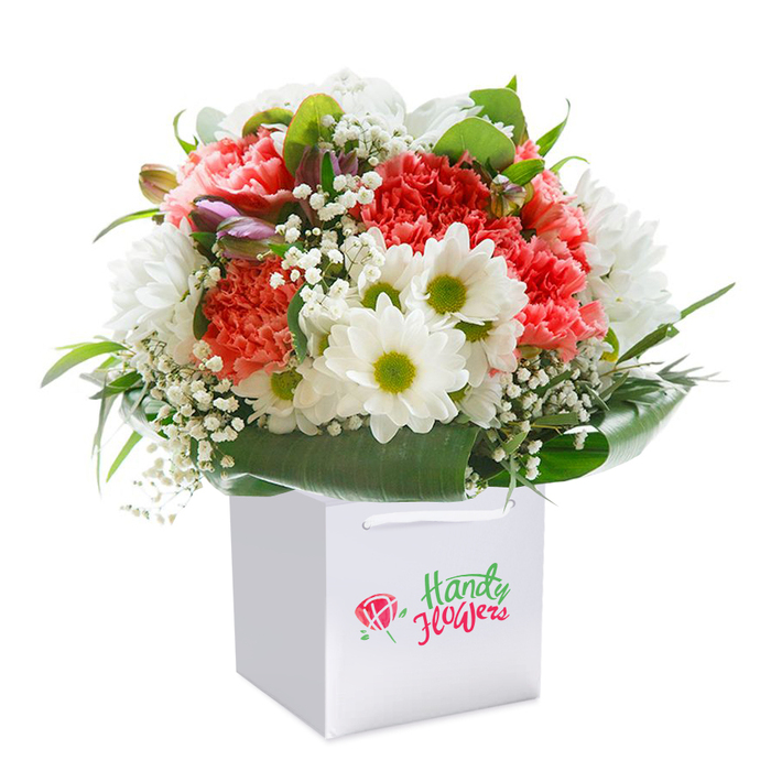 Set against a flawless white background, this lush, expertly arranged bouquet stands proudly in a white, square gift bag-a picture of modern Purley elegance. The display draws the eye with full coral and peach carnations, their distinctive ruffled petals offering a sumptuous visual and sensory feast. Bright white daisies with lemon-lime centers bring an uplifting brightness, echoing the freshness of green spaces like Purley Way Playing Fields. Wisps of delicate baby's breath trace through the arrangement, creating gentle, airy layers that soften the bouquet's texture. Scattered on the left, closed buds with a hint of purple intrigue the viewer, alluding to the evolving gardens along Brighton Road. Everything is embraced by deep, glossy green leaves, cradling the base and shaping the bouquet into a generous, rounded silhouette that promises both quality and abundance. The accompanying white gift bag, complete with white ribbon handles, is designed for effortless delivery throughout Purley-whether sent as an anniversary gesture, birthday delight, or spontaneous reminder of care. Every element, from the selection of flowers to the careful arrangement, reflects the skill and dedication typical of Purley's beloved local florists, ready to bring a moment of joy and freshness to any doorstep.