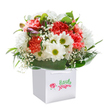 A burst of colour and vivacity-this floral bouquet, upright in a chic white square gift bag, channels the cheerful spirit of Purley's high streets and green corners. Coral carnations with deeply textured, ruffled petals mingle with peach tones, exuding a sense of warmth and comfort. Crisp, white daisies crowned with yellow-green centers lift the mood, much like a walk through the gardens of Oakwood Park. Wisps of baby's breath thread delicately between the larger blooms, creating a playful lightness. Peeking from the left, tender purple-tinged buds draw the eye, adding a layer of gentle intrigue that hints at new beginnings. Surrounding the floral centerpiece is a collar of deep, glossy green foliage, framing the arrangement and providing vivid depth. The ensemble is thoughtfully nestled in a sturdy white gift bag with ribbon handles, ready to surprise a loved one for a birthday, anniversary, or heartfelt 'thinking of you' moment anywhere in Purley. The attention to artistry and freshness shines-each flower reflecting the genuine warmth and community focus of local Purley florists, ready to deliver beauty and happiness neighbourhood-wide.